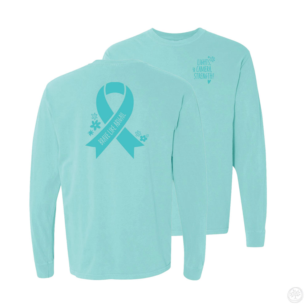 Light blue long sleeve shirt with large teal awareness ribbon on back reading “Brave Like Abigail,” and front text that says “Lights, Camera, Strength!” – custom childhood illness awareness apparel.