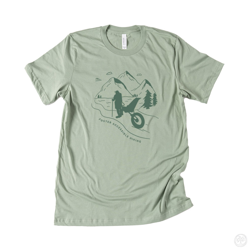 Sage green short sleeve t-shirt with mountain scene and silhouette of wheelchair hiker, reading “Foster Accessible Hiking” – awareness design for accessibility in nature and inclusion.