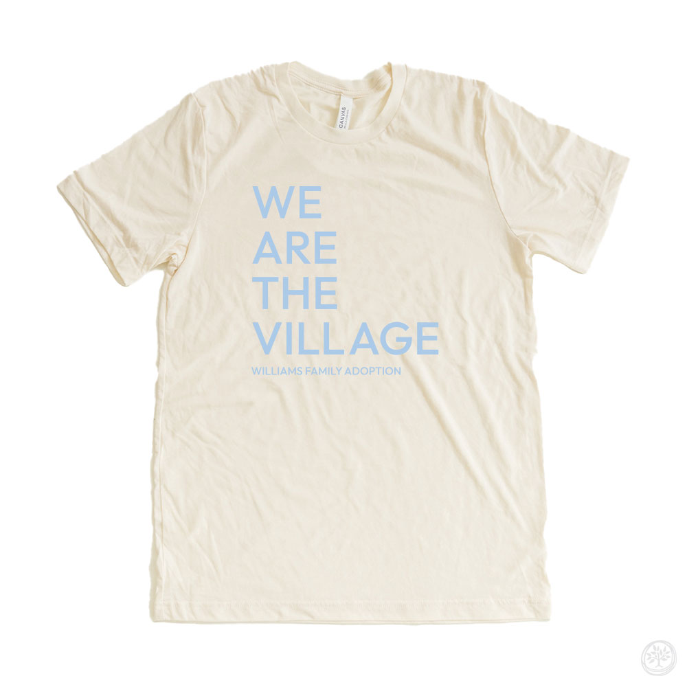 "WE ARE THE VILLAGE" tee (ivory): Ivory t-shirt with light blue text that says "WE ARE THE VILLAGE – Williams Family Adoption," in a modern, stacked layout.