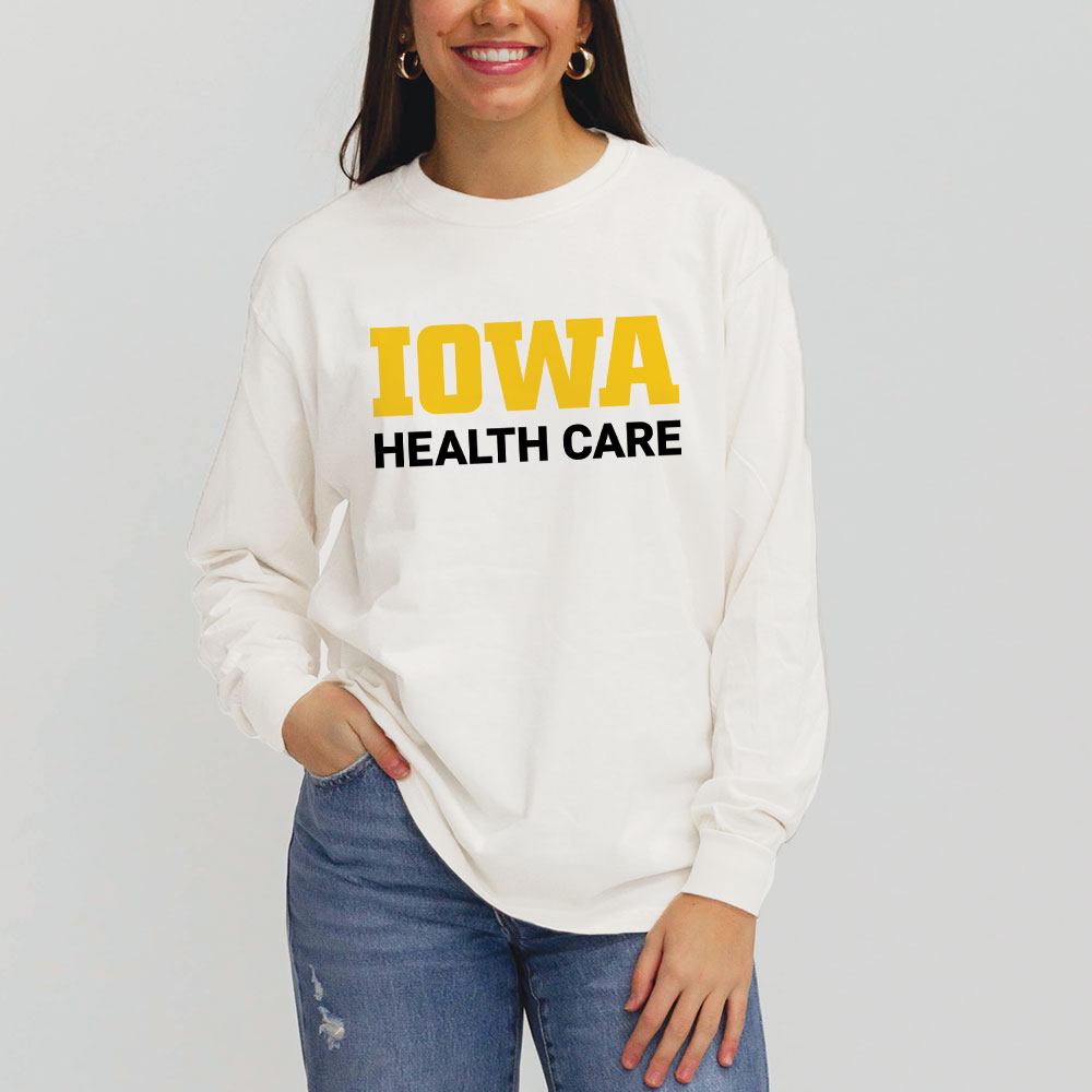 A woman is smiling and wearing a white long-sleeve t-shirt with the words “IOWA” in bold yellow letters and “HEALTH CARE” in black capital letters underneath. She is casually posing with one hand in her pocket, wearing ripped blue jeans. The background is