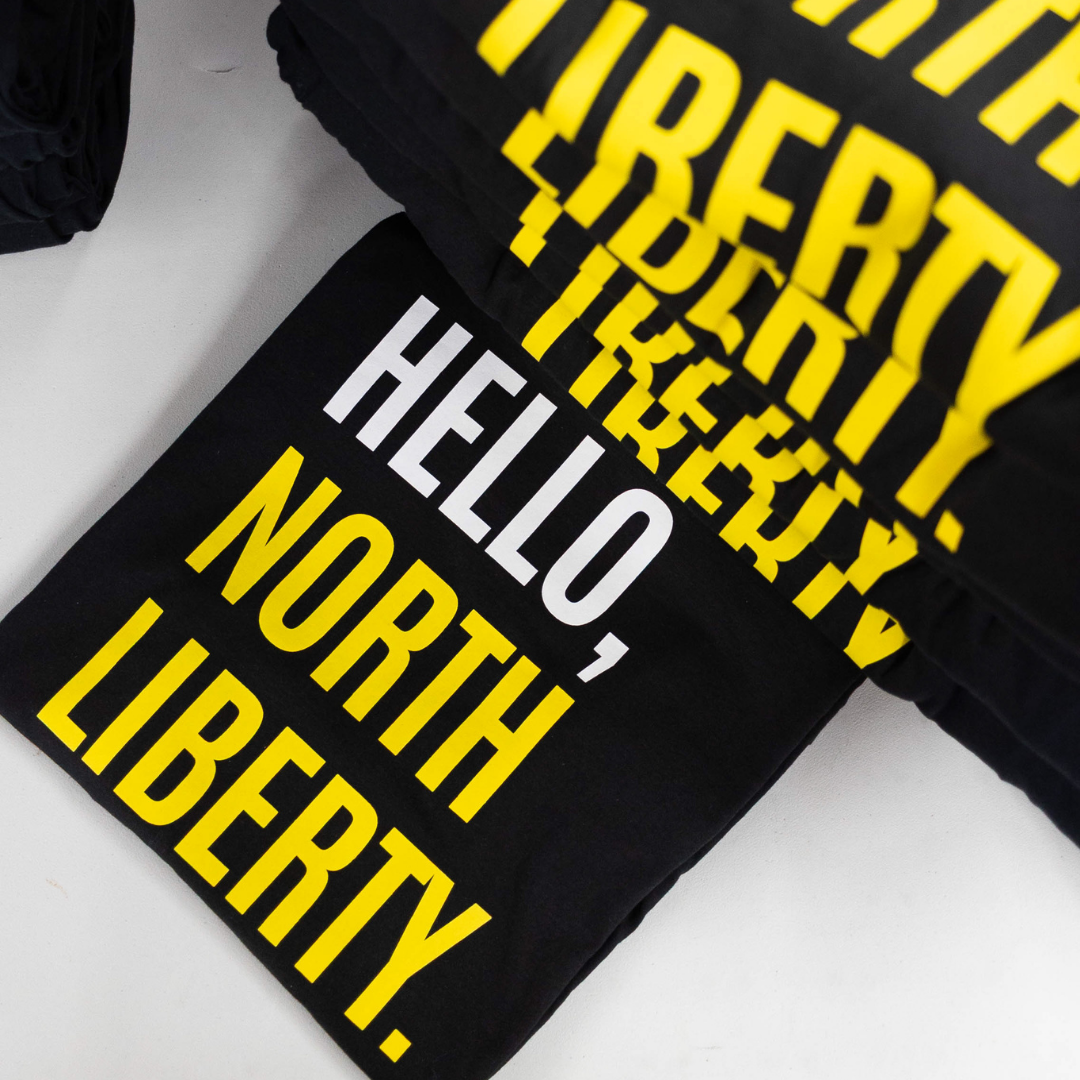 Stacked black t-shirts with bold yellow and white text reading “Hello, North Liberty,” screen printed for a bulk order
